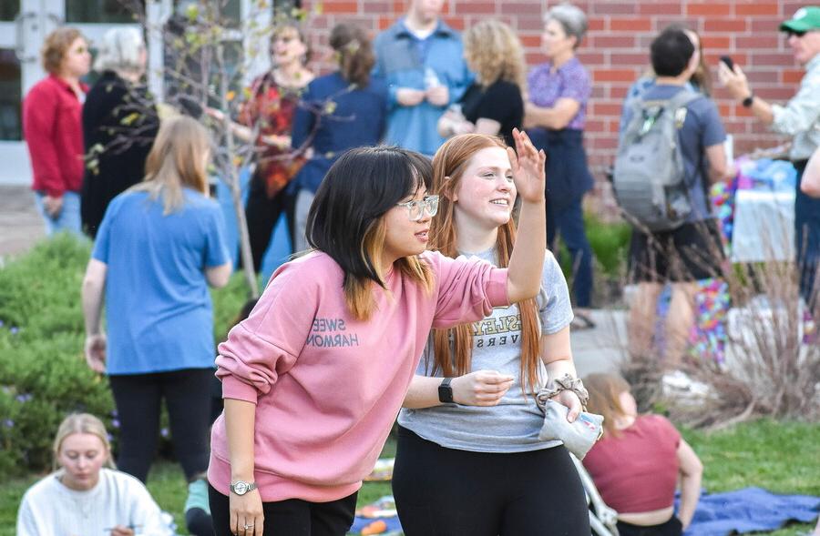 Two young women tossing a sand bag amongst many people outdoors.